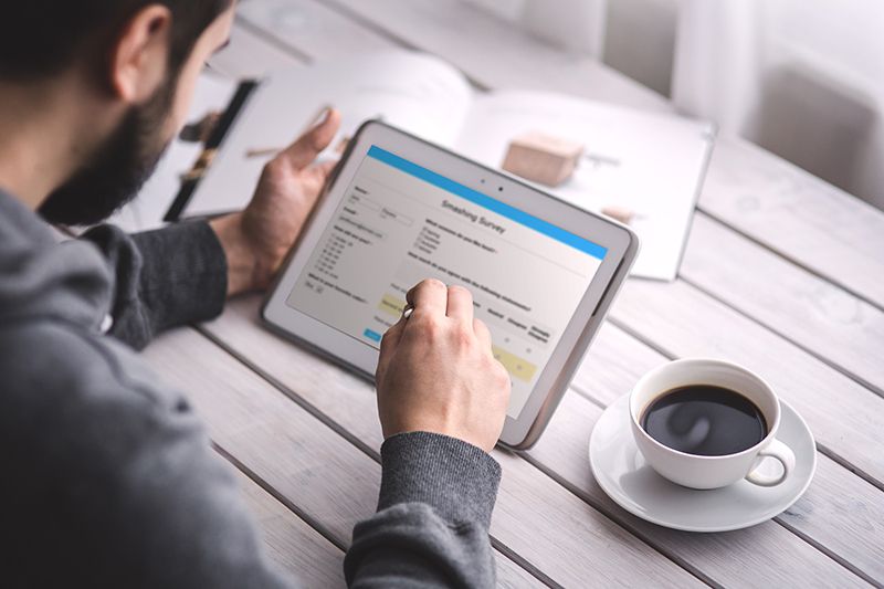 Photo of Man at Table With Tablet in Hand Completing Survey Coffee on Table and Book